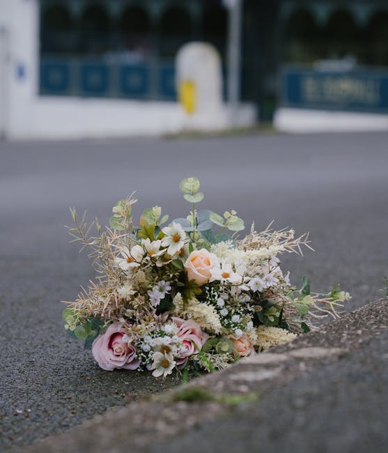 A floral bouquet featuring soft pastel-colored roses, white daisies, and delicate filler flowers arranged with eucalyptus and other greenery, resting on a paved surface near a curb. The arrangement is
