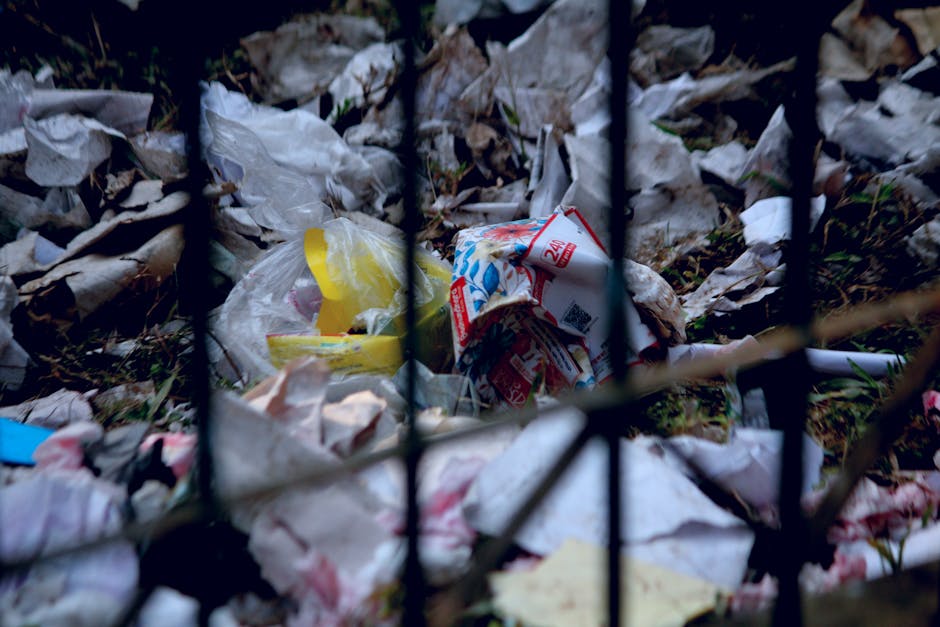 A close-up view of a pile of discarded trash behind a black metal fence, including crumpled paper, plastic wrappers, and a flattened cardboard box, set against a background of dried grass and leaves.