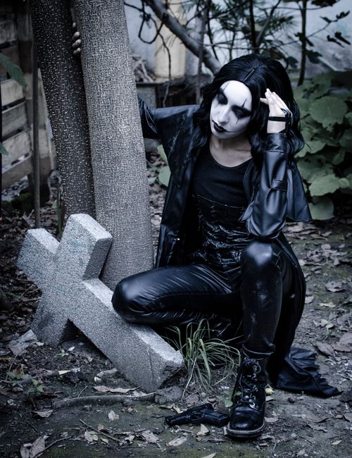 A person with pale makeup, dark hair, and gothic-style black clothing, including a leather jacket and pants, is sitting outdoors next to a weathered wooden post and a small stone cross. They are crouc