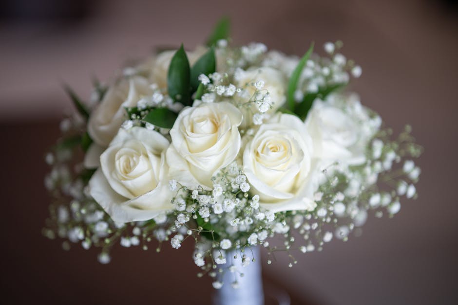 A close-up image of a floral bouquet featuring creamy white roses with lush, velvety petals arranged in a rounded formation, complemented by sprigs of delicate white baby's breath flowers with tiny cl