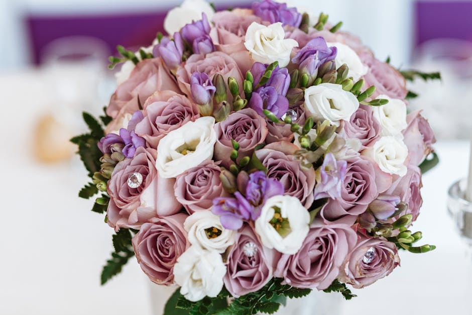 A close-up of a floral bouquet featuring pastel pink roses, white lisianthus, and purple freesia arranged in a rounded, densely packed style. The flowers are fresh, with slightly ruffled petals and gr