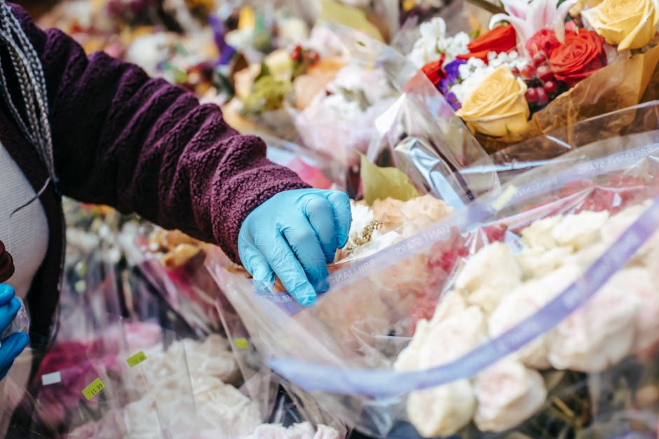 A person wearing a dark purple textured long sleeve top and blue gloves is carefully selecting and arranging flowers from a large display of various fresh floral bouquets. The flowers include cream, r