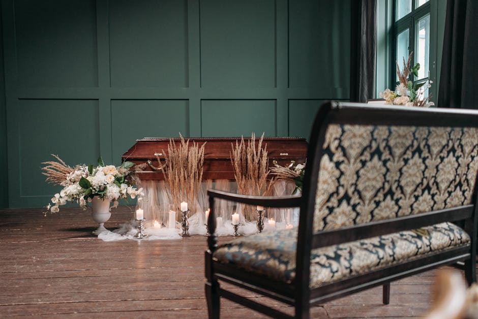 A wooden funeral casket is decorated with a floral arrangement consisting of white roses, baby's breath, and lush green foliage, placed against a deep green paneled wall in a softly lit room. Surround