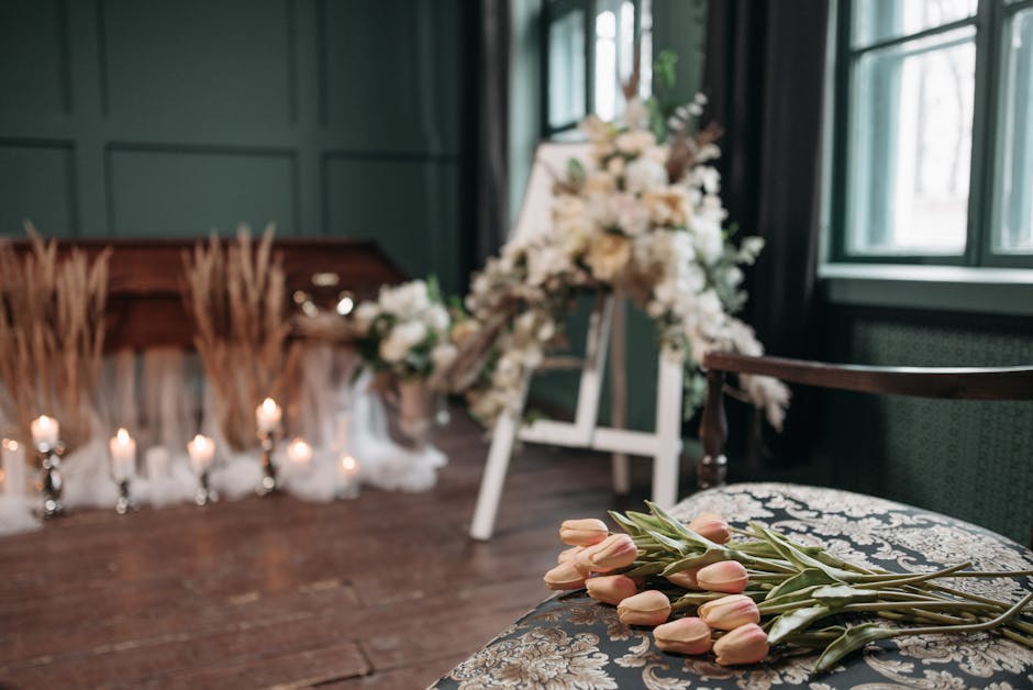 A floral arrangement scene featuring a bouquet of pale pink tulips with green stems and leaves resting on a patterned fabric surface. In the background, there is a standing easel decorated with a larg