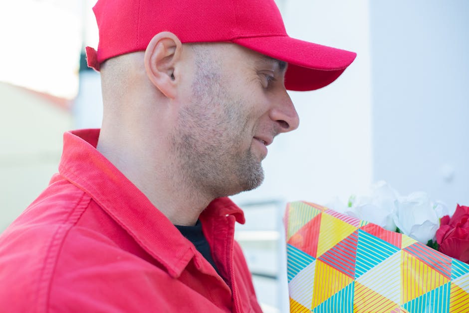 A man dressed in a red jacket and matching red cap holds a brightly wrapped floral gift box with a colorful geometric pattern. The bouquet inside the box features vibrant pink and white flowers, with 