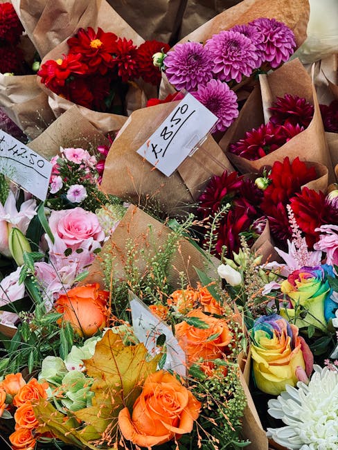 A colorful assortment of freshly cut flowers arranged in brown paper wrapping, showing vibrant red dahlias, purple chrysanthemums, soft pink roses, and rainbow-hued roses, all with lush green foliage